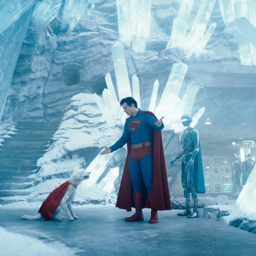 Dressed in his blue and red suit, Superman stands in a cave made of ice. Next to him is a white dog wearing a matching red cape.